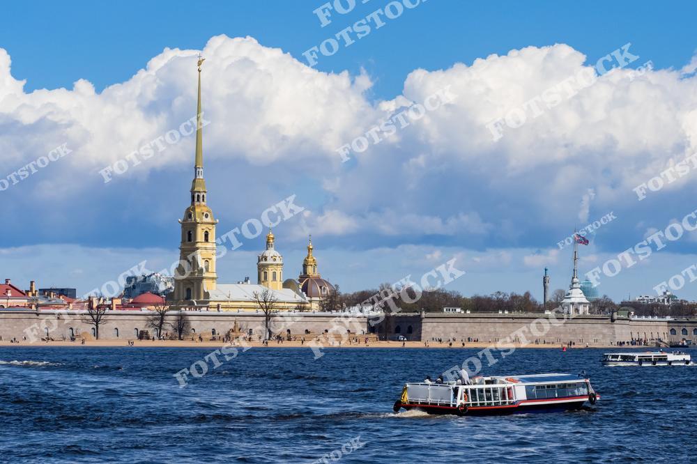 На фотографии изображена Петропавловская крепость, один из знаковых символов Санкт-Петербурга, расположенная на Заячьем острове. Золотой шпиль Петропавловского собора возвышается над крепостью, придавая композиции величественный вид. На переднем плане виден прогулочный катер, курсирующий по Неве, добавляющий динамику в кадр. Солнечная погода и яркие облака делают снимок особенно живописным.